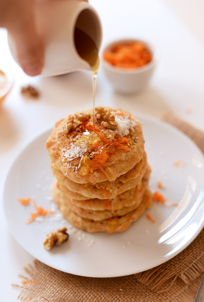 Pouring syrup onto a stack of Carrot Cake Pancakes made with coconut and walnuts
