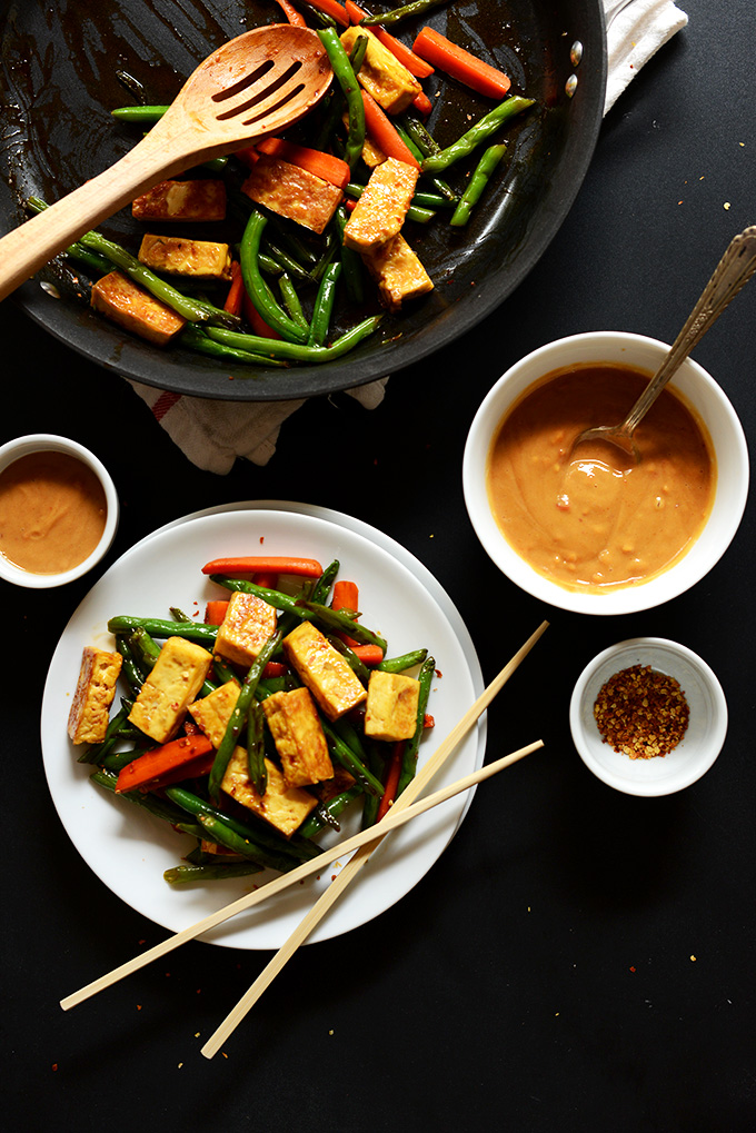 Plate and skillet of Tofu Stir Fry with bowls of peanut sauce and red chili flakes