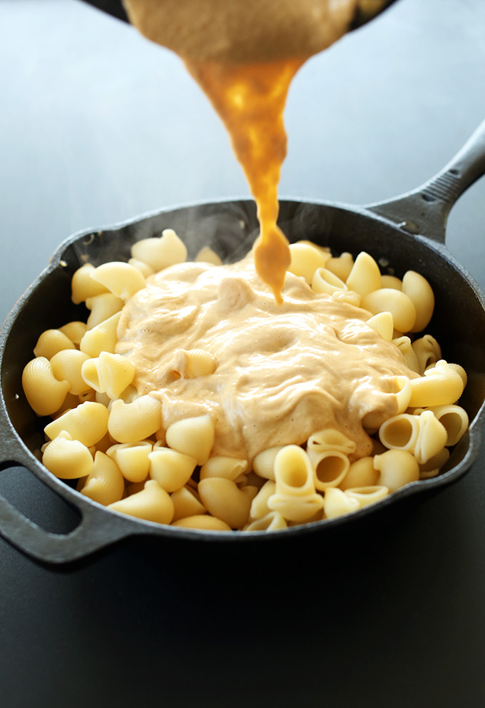 Pouring vegan cheese sauce onto skillet of macaroni noodles