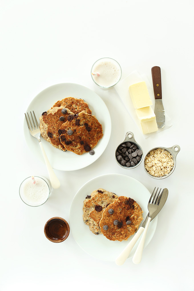 Two plates of GF Chocolate Chip Oatmeal Cookie Pancakes surrounded by ingredients used to make them