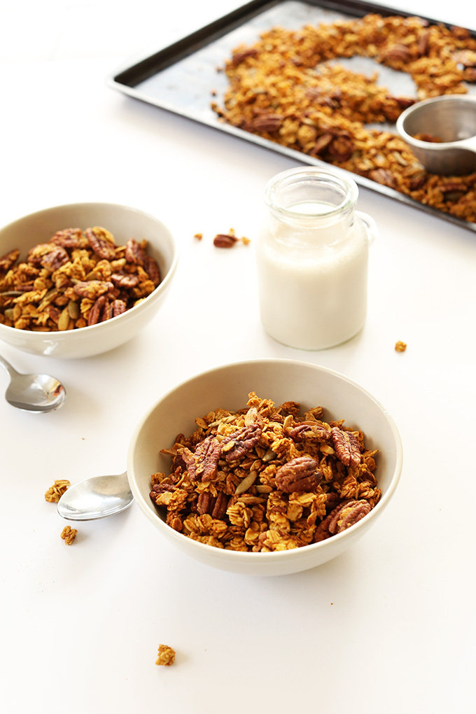 Serving bowls and baking sheet filled with Pumpkin Pecan Granola alongside a glass of nut milk