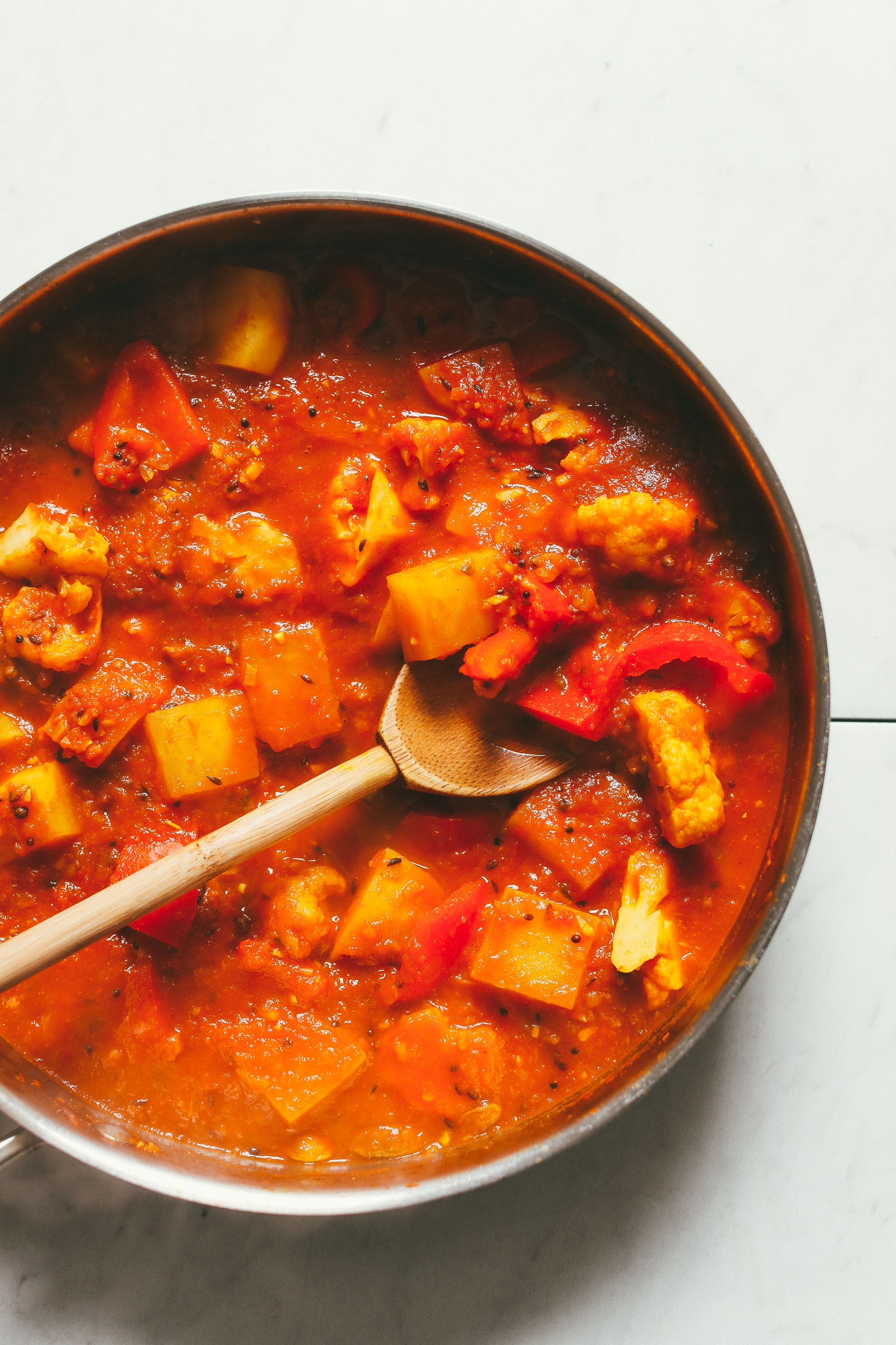 An overhead image of a pan with red lentil curry, potatoes, red bell pepper, and cauliflower being stirred with a wooden spoon