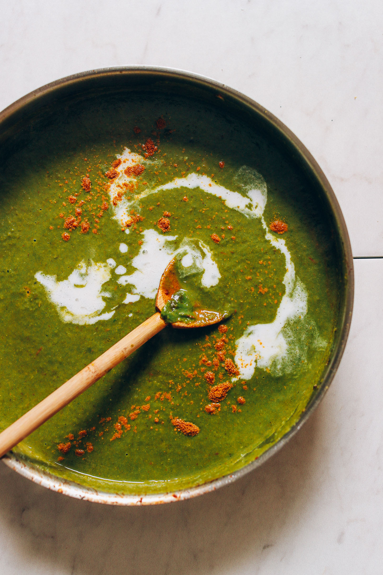Overhead image of a pan with palak paneer sauce being stirred with wooden spoon, garnished with curry power and coconut milk