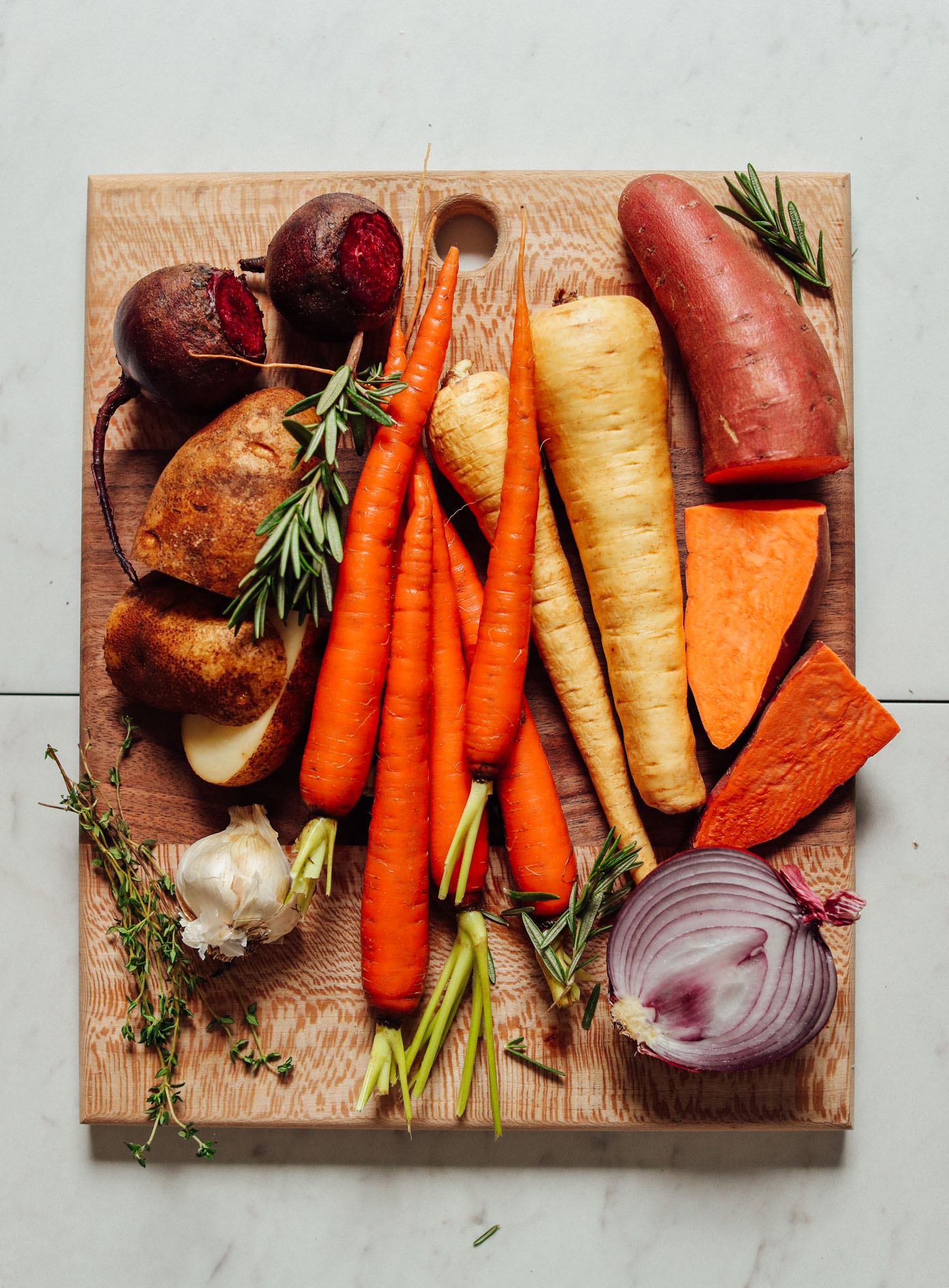 Wood cutting board filled with fresh beets, potatoes, parsnips, carrots, onions, garlic, and herbs for roasting