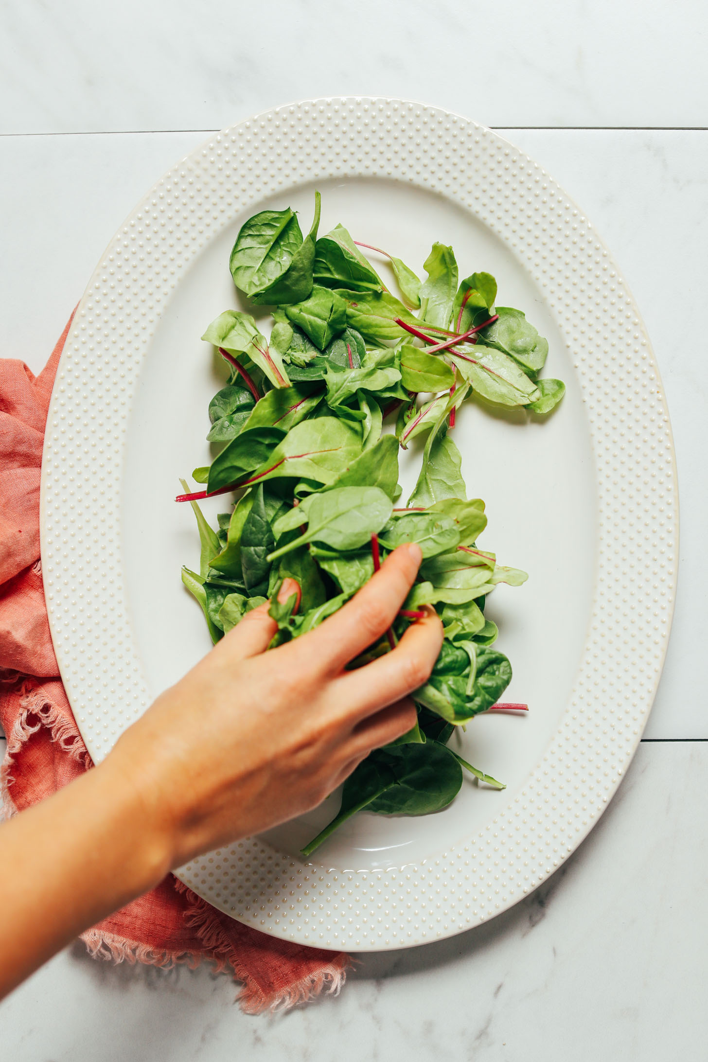 Overhead image of a white platter pink towel and greens being placed on the plate by hand