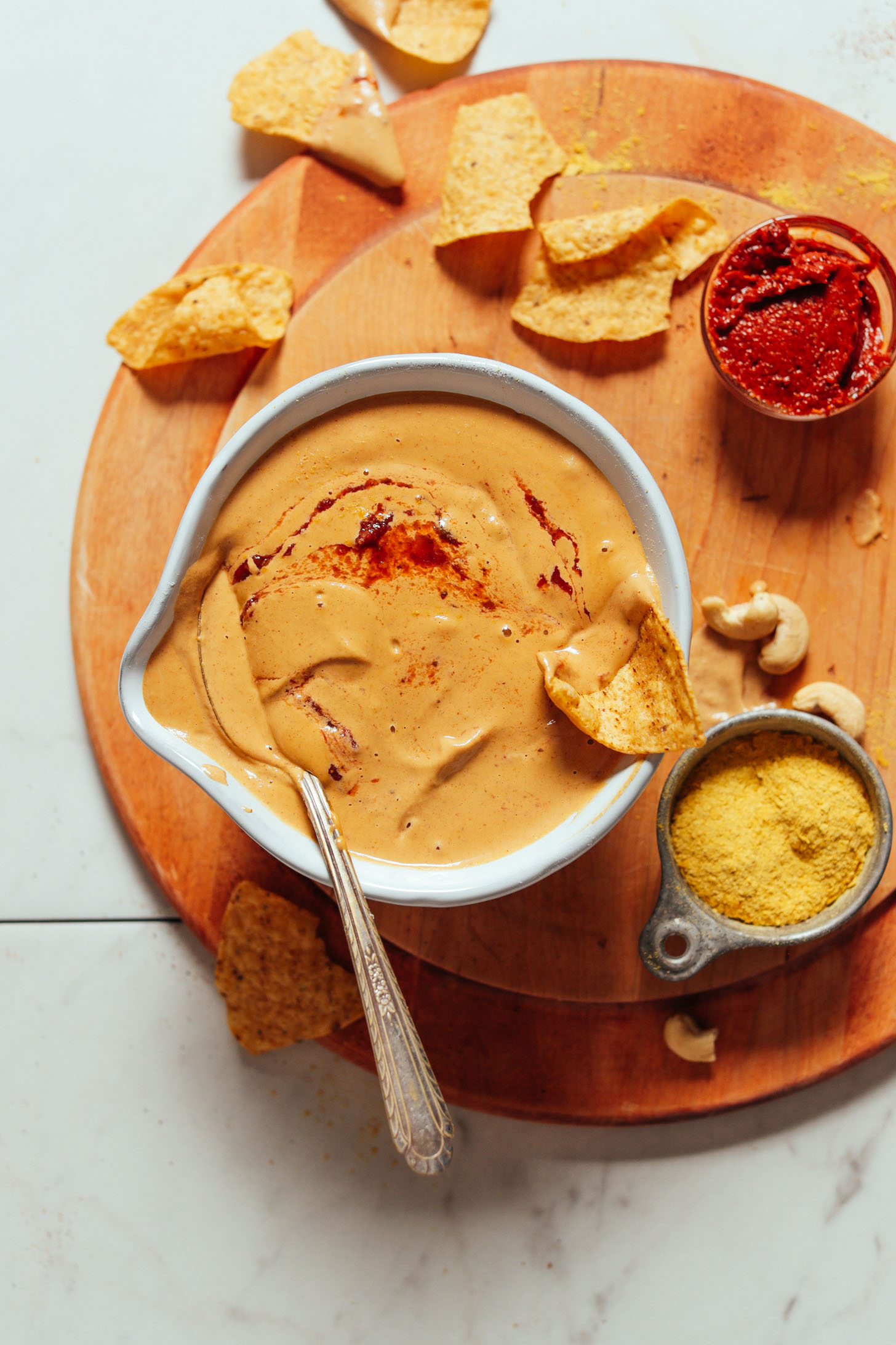 Overhead image of vegan cashew queso in a white bowl on a cutting board with harissa, chips, and nutritional yeast on the side