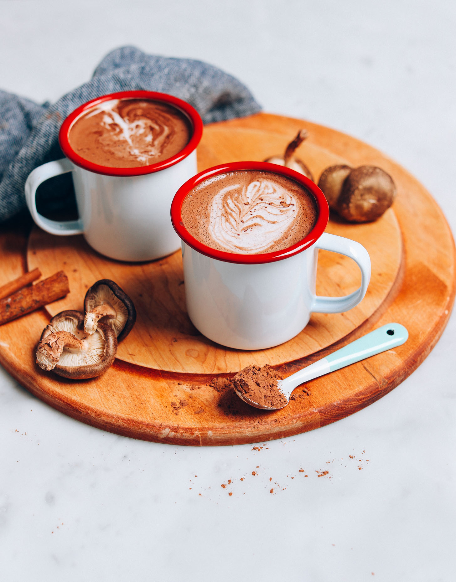 Straight on image of mushroom cacao hot latte on a cutting board with a blue towel, mushrooms in the background, and blue spoon of cacao powder in front