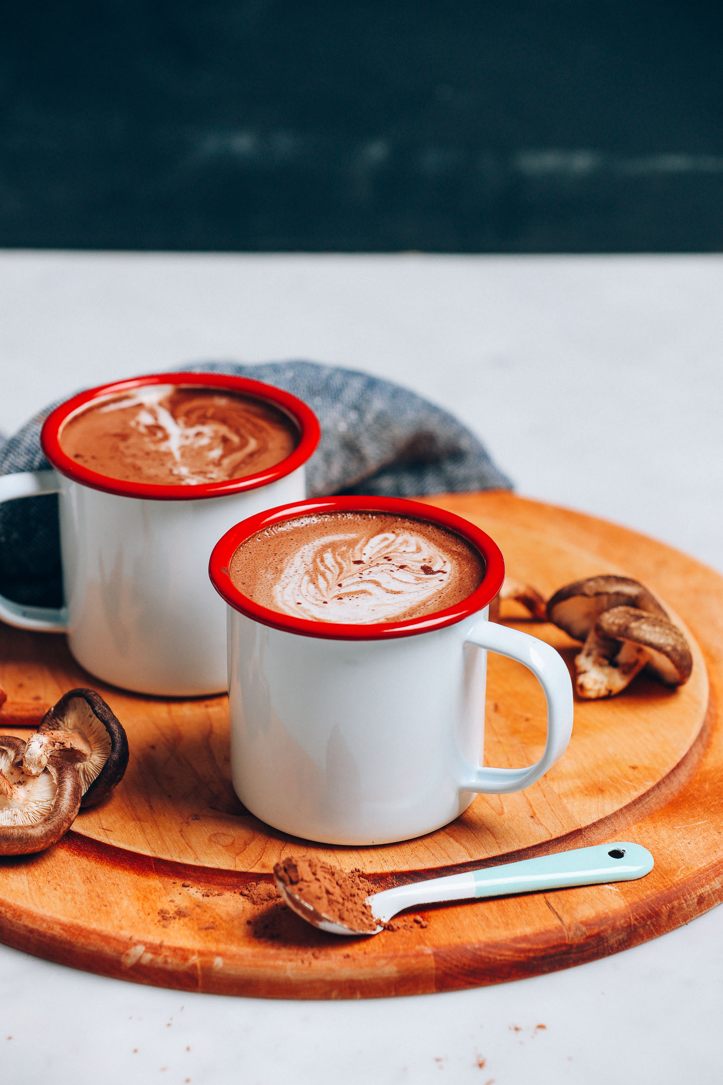 Straight on image of hot cacao mushroom latte on a cutting board with blue towel, mushrooms on the side, and a blue spoon with cacao powder in the front