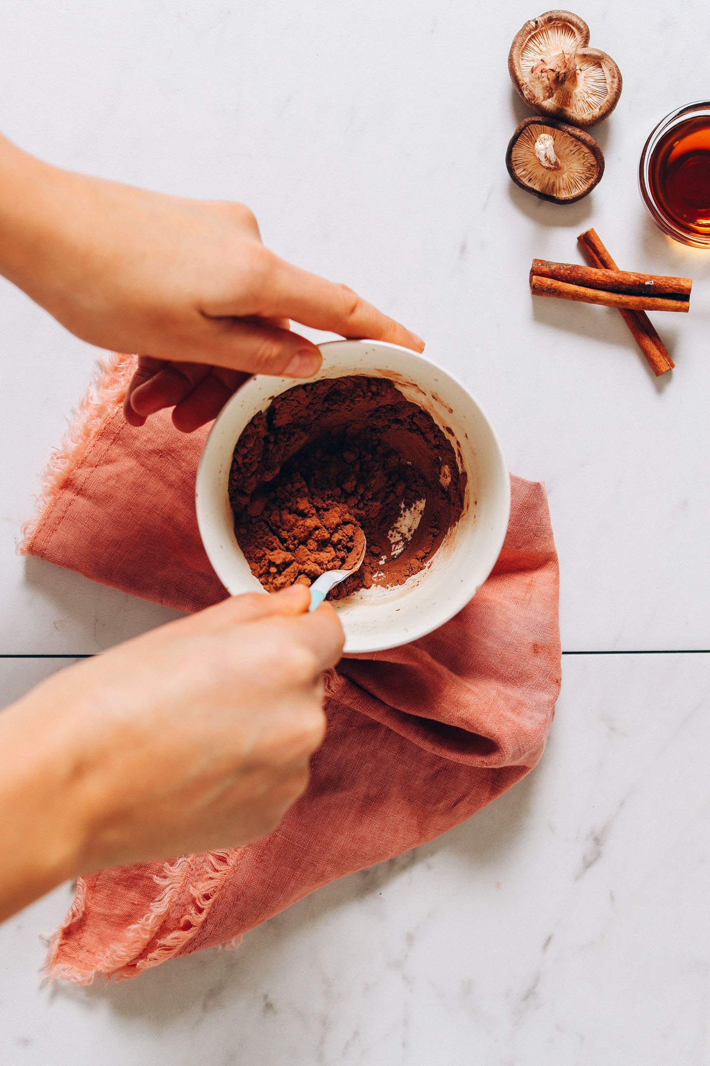 Overhead image of white bowl on a pink towel and a hand stirring cacao powder