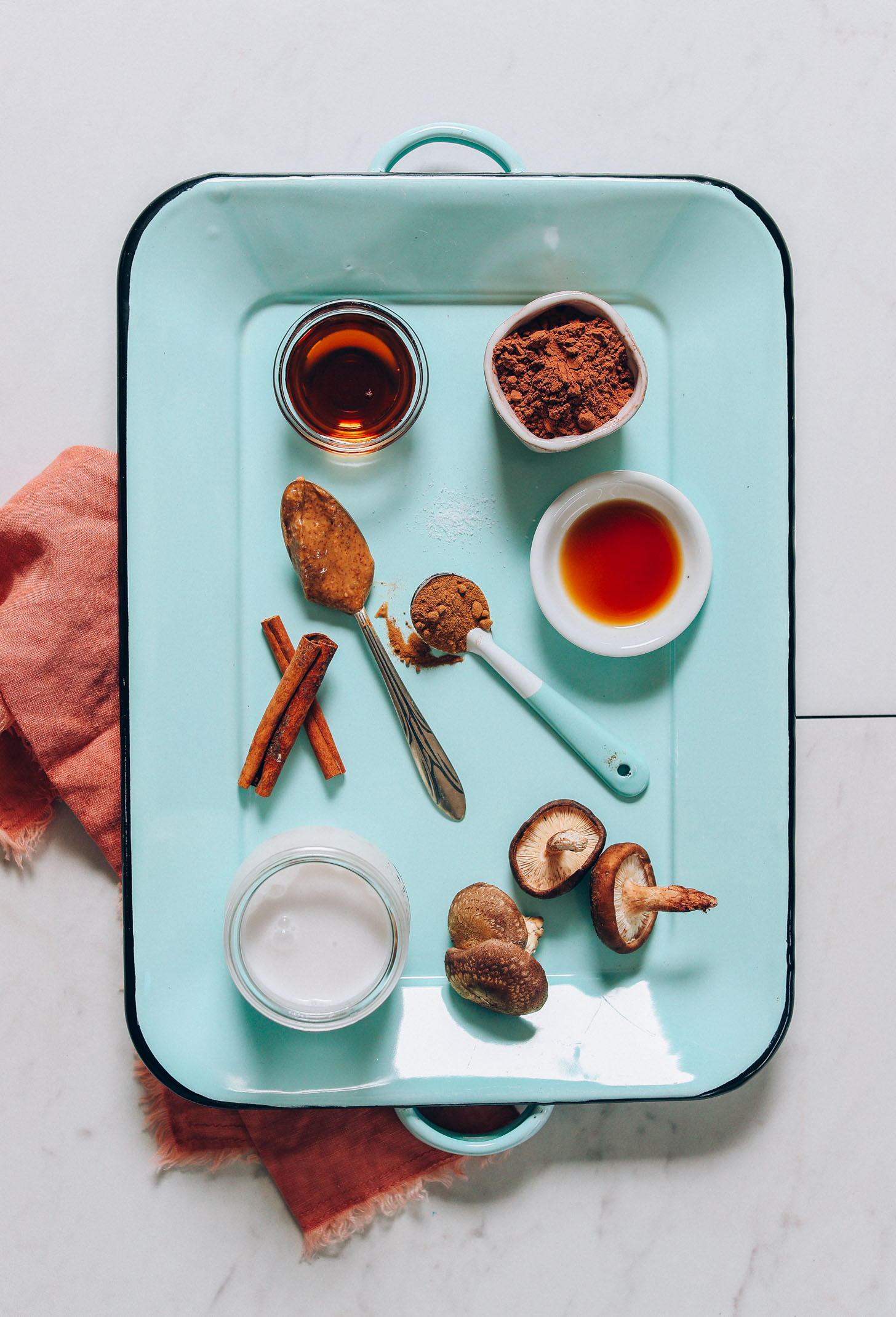 Overhead image of blue tray holding ingredients for mushroom hot cacao latte including mushroom powder, maple syrup, cinnamon, and coconut milk