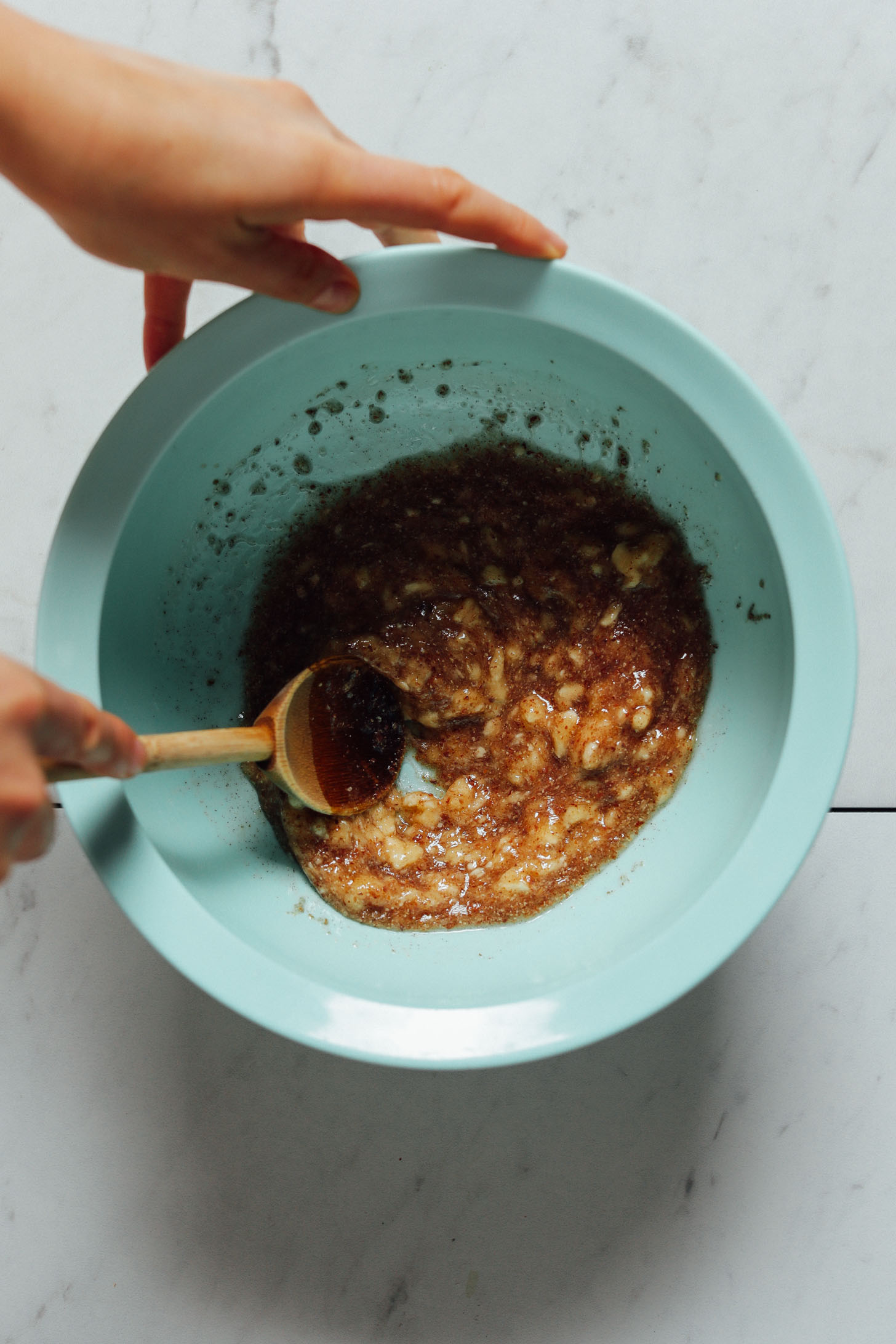 Overhead image of ripe banana mixed with flax seed, maple syrup, and oil in a blue bowl with a wooden spoon stirring the mixture