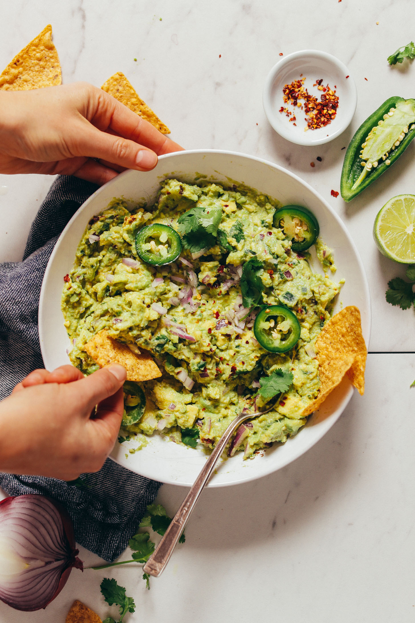 Overhead image of guacamole in a white bowl garnished with fresh jalapenos, and a hand with a chip scooping some guacamole