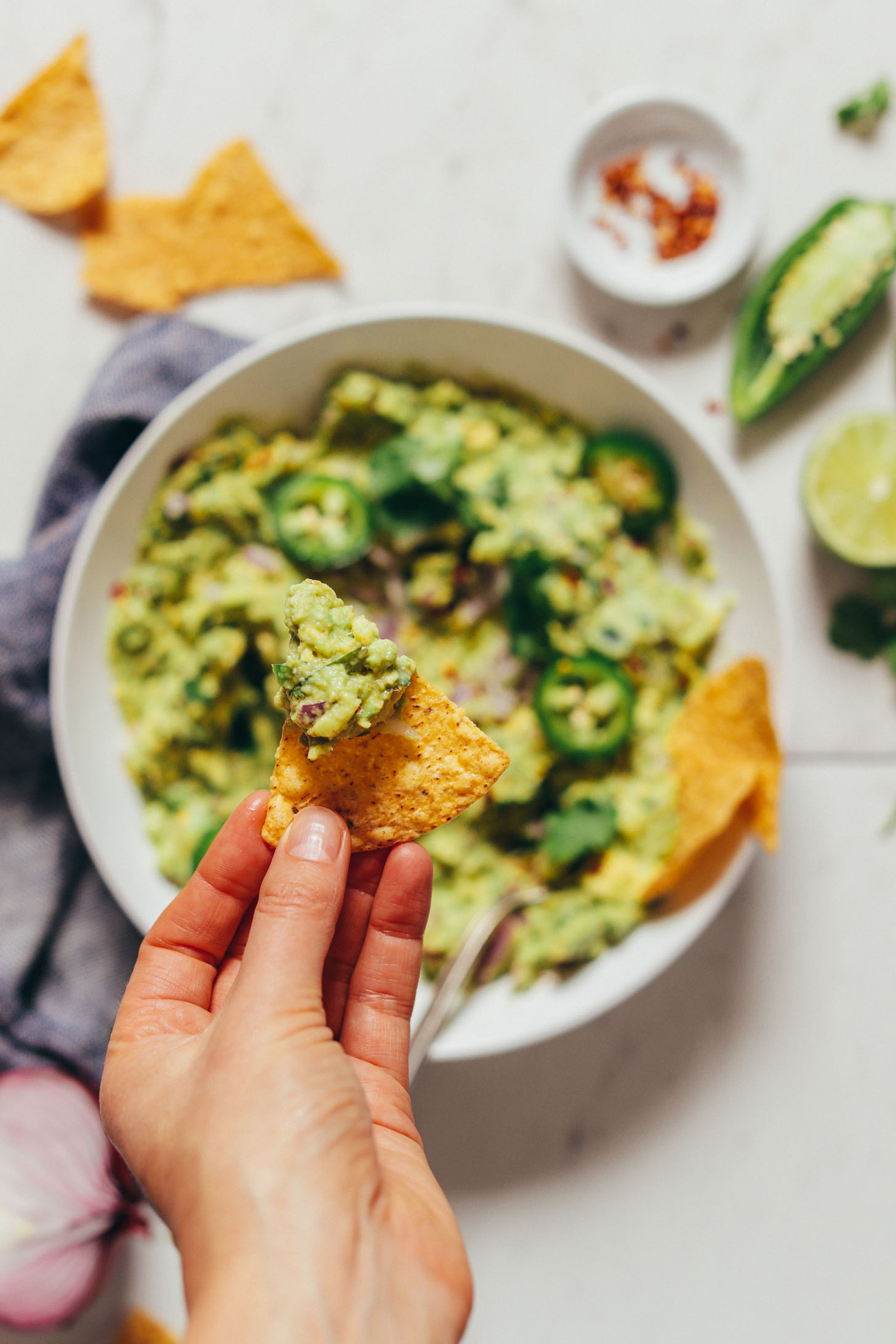 An overhead image of guacamole in a white bowl in the background with a hand holding a chip with some guacamole in focus overhead