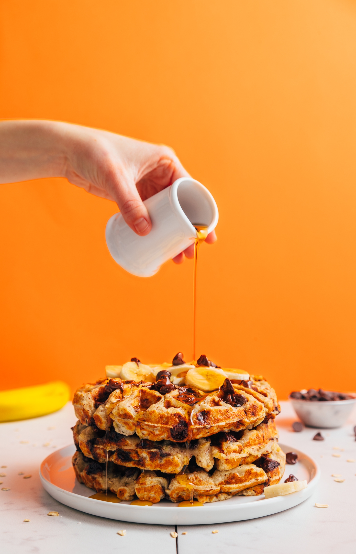 Drizzling maple syrup onto a stack of Chocolate Chip Banana Bread Waffles
