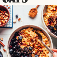 Hands holding the side of a bowl and a spoon in a bowl of creamy pumpkin oats with blueberries and almonds