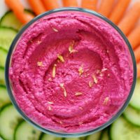 Bowl of vibrant pink Roasted Beet Hummus surrounded by carrot and cucumber slices