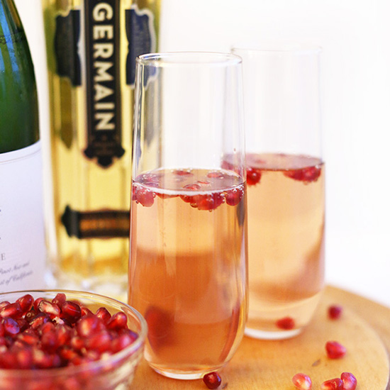 Cutting board with a bowl of pomegranate arils beside fancy glasses filled with our St Germain Spritzers recipe