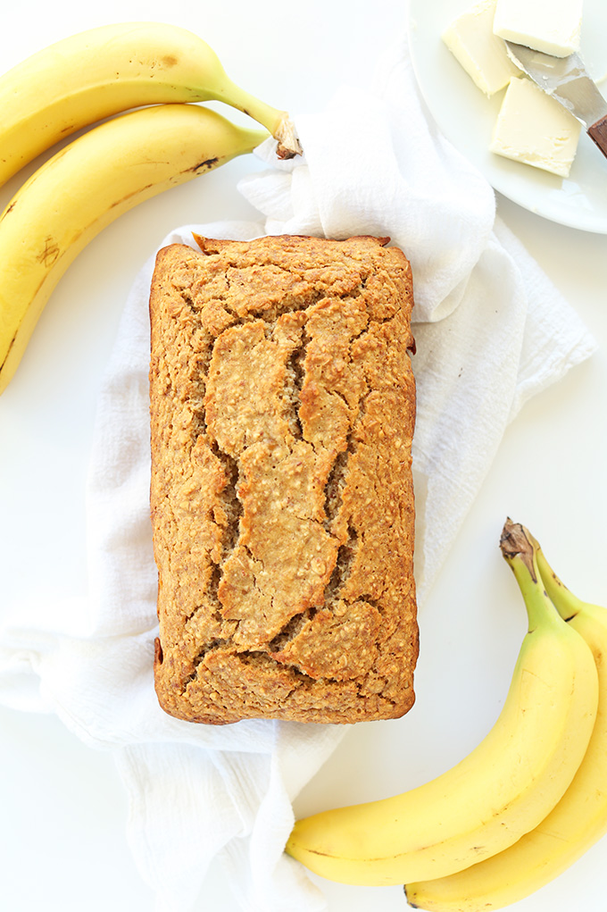 Freshly baked loaf of simple Gluten Free Banana Bread surrounded by fresh bananas and vegan butter
