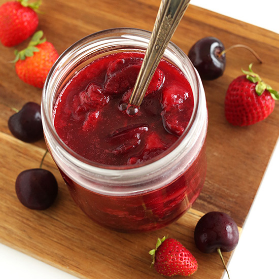 Spoon in a jar of Simple Berry Compote beside fresh strawberries and cherries