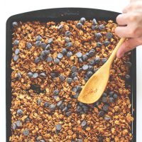 Using a wooden spoon to stir a batch of Peanut Butter Chocolate Chip Granola on a baking sheet