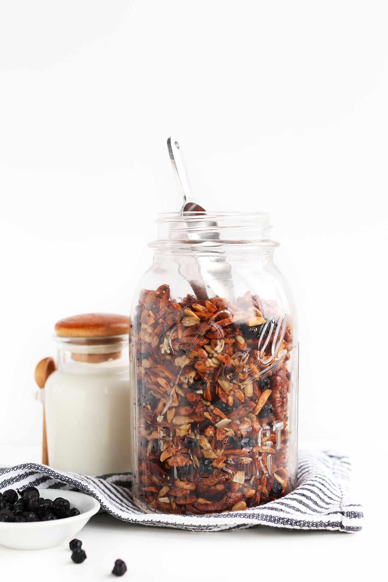 Jar of simple homemade vegan Grain-Free Granola alongside nut milk and dried blueberries