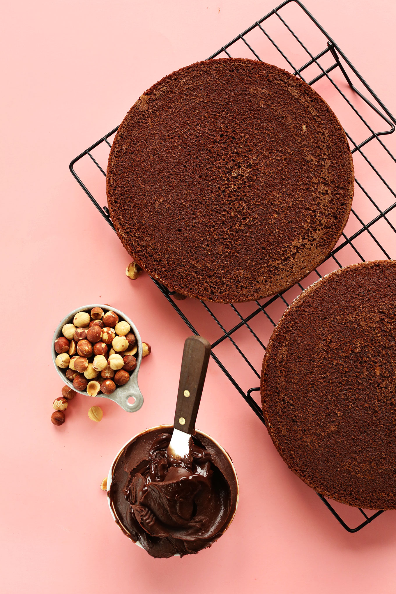 Rich and moist Chocolate Hazelnut Cakes on a cooling rack beside frosting and roasted hazelnuts for decorating