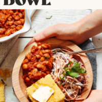 Hands holding the sides of a vegan BBQ bowl filled with beans, coleslaw, and cornbread