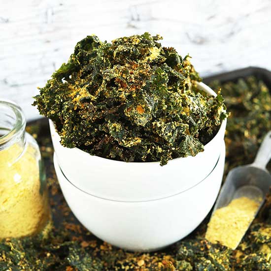 Bowl of Cheesy Kale Chips on a baking sheet with more chips and nutritional yeast