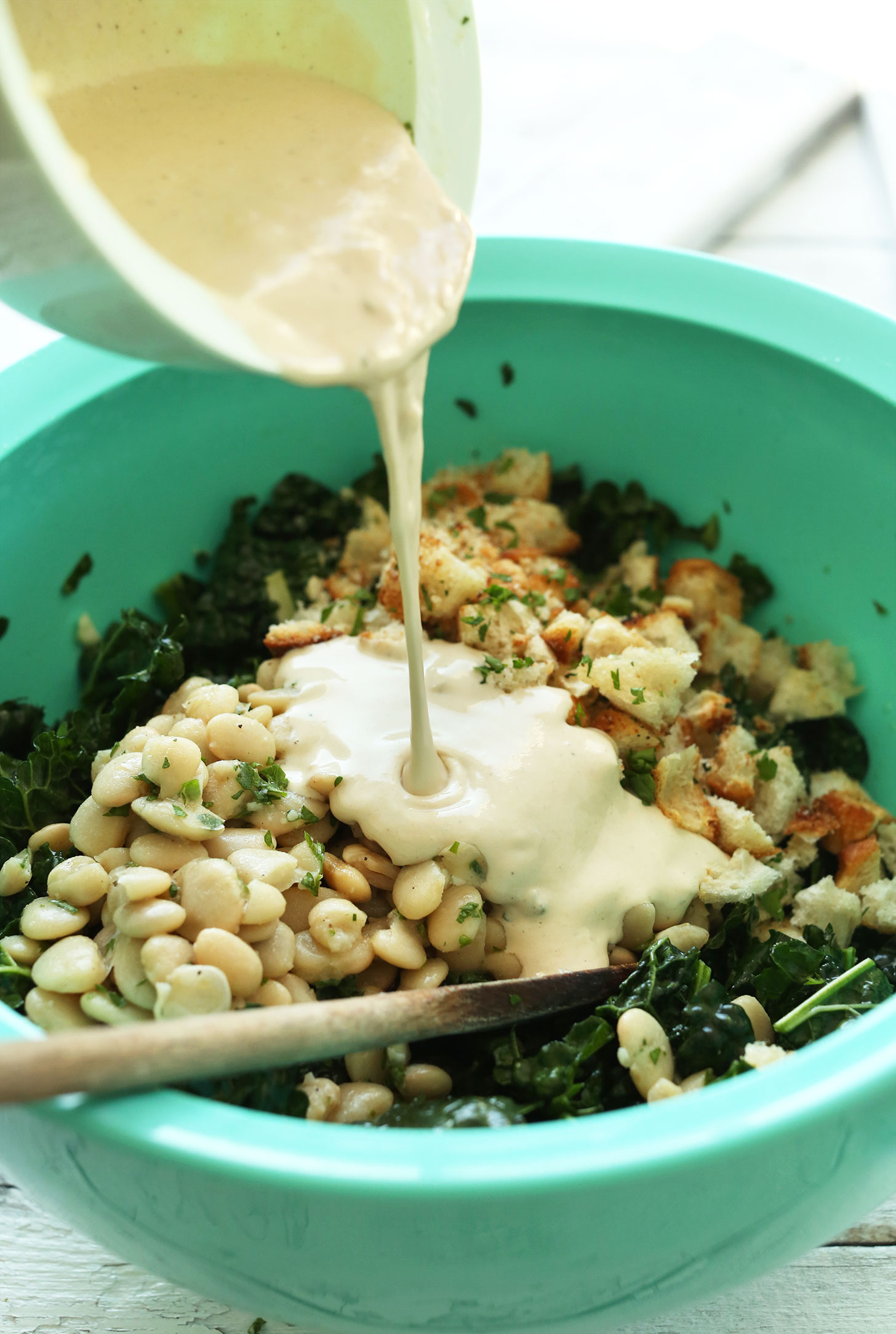 Pouring Garlic-Tahini Dressing over our Lemony Garlic Kale Salad with Butter Beans