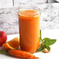 Tall jar of Carrot Apple Ginger Juice surrounded by ingredients used to make it