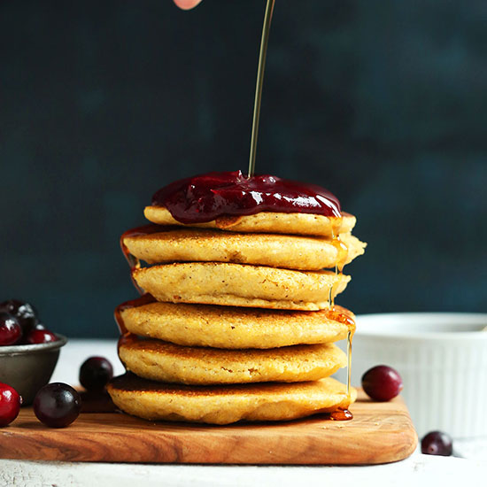 Drizzling syrup onto a stack of Cornmeal Pancakes topped with Cranberry Compote