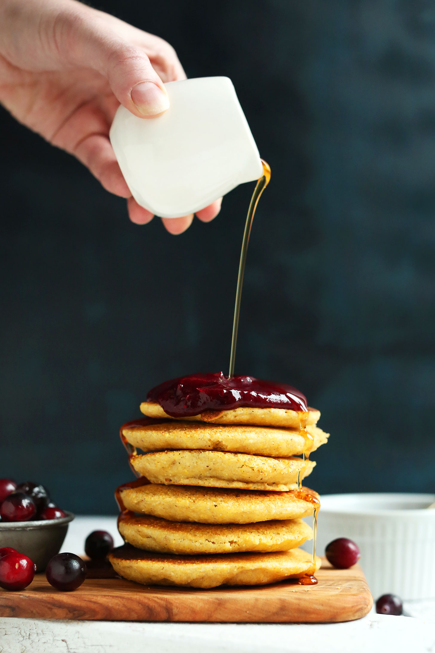 Drizzling syrup onto a stack of gluten-free vegan cornmeal pancakes topped with cranberry compote