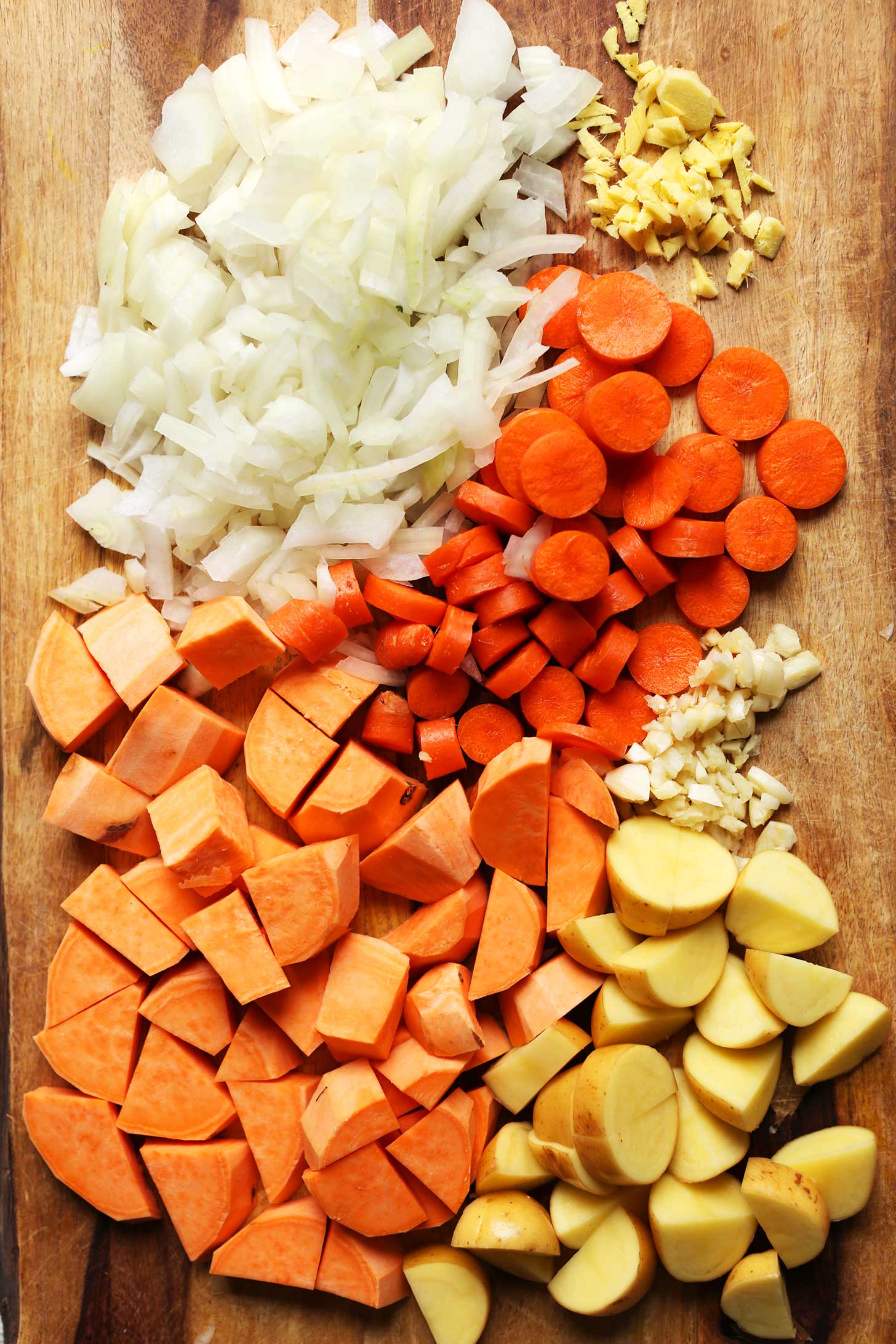 Wood cutting board with onions, carrots, garlic, ginger, sweet potatoes, and potatoes for making healthy gluten-free vegan soup