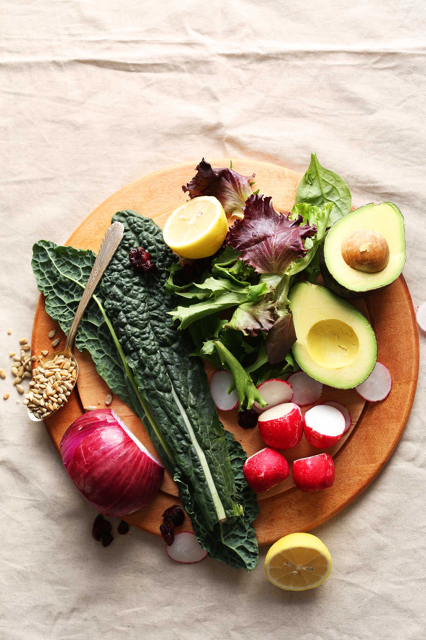 Wood cutting board featuring kale, red onion, radishes, avocado, lemon, sunflower seeds, cranberries, and greens for a delicious plant-based salad recipe