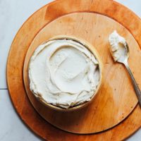 Spoon and bowl of Cashew Buttercream Frosting on a wood cutting board