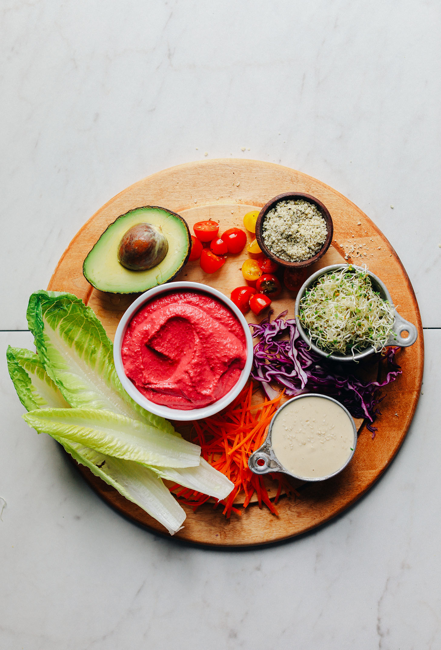 Wood cutting board displaying romaine lettuce boats, carrots, red cabbage, tomatoes, alfalfa sprouts, hemp seeds, avocado, beet hummus, and tahini dressing for Raw Romaine Taco Boats