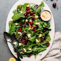 Big platter of Mustard Green Cherry Salad with a measuring cup filled with tahini dressing