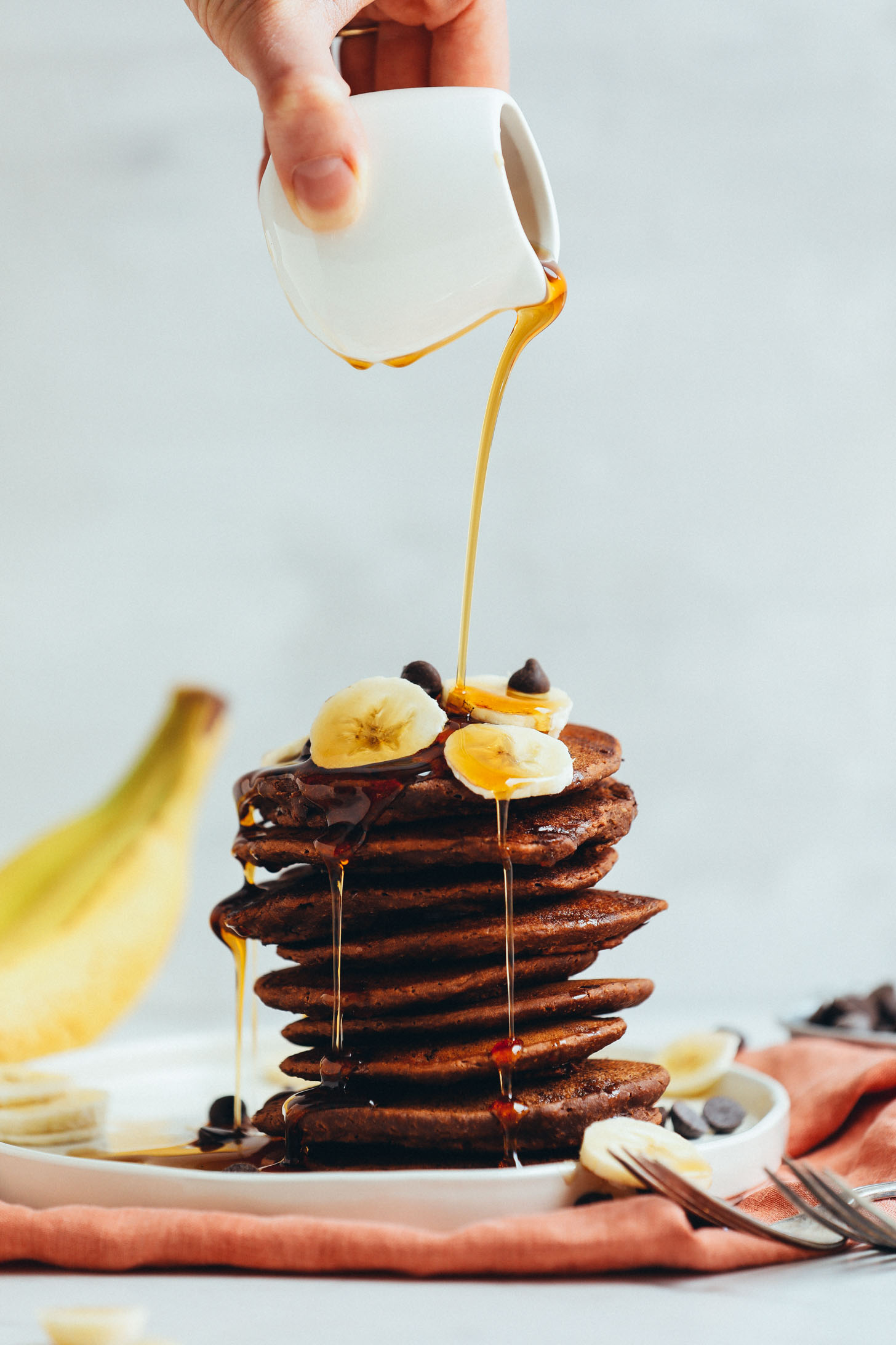 Pouring maple syrup onto a stack of naturally-sweetened Vegan Chocolate Chocolate Chip Pancakes