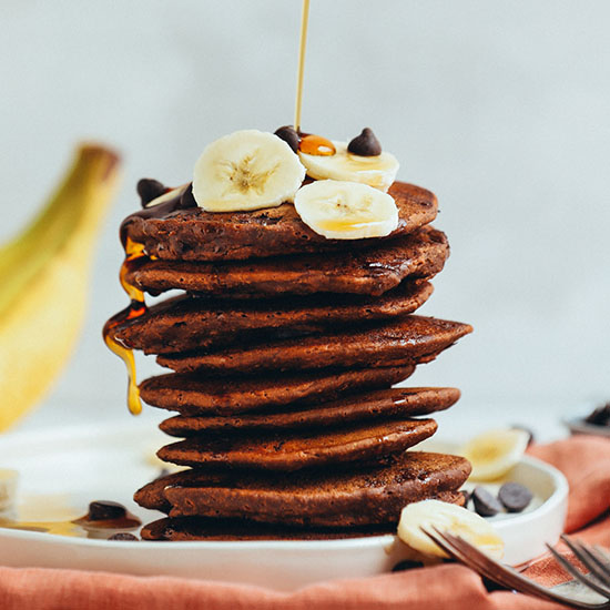 Drizzling syrup onto a stack of Chocolate Chocolate Chip Pancakes topped with sliced banana