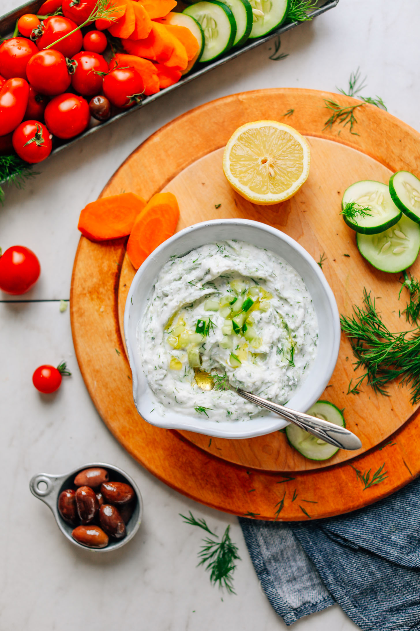 Cutting board with a bowl of Vegan Tzatziki surrounded by key ingredients and veggies for dipping