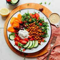 Plate filled with our Easy Greek Bowl recipe surrounded by tahini sauce, lemon, and sliced vegetables
