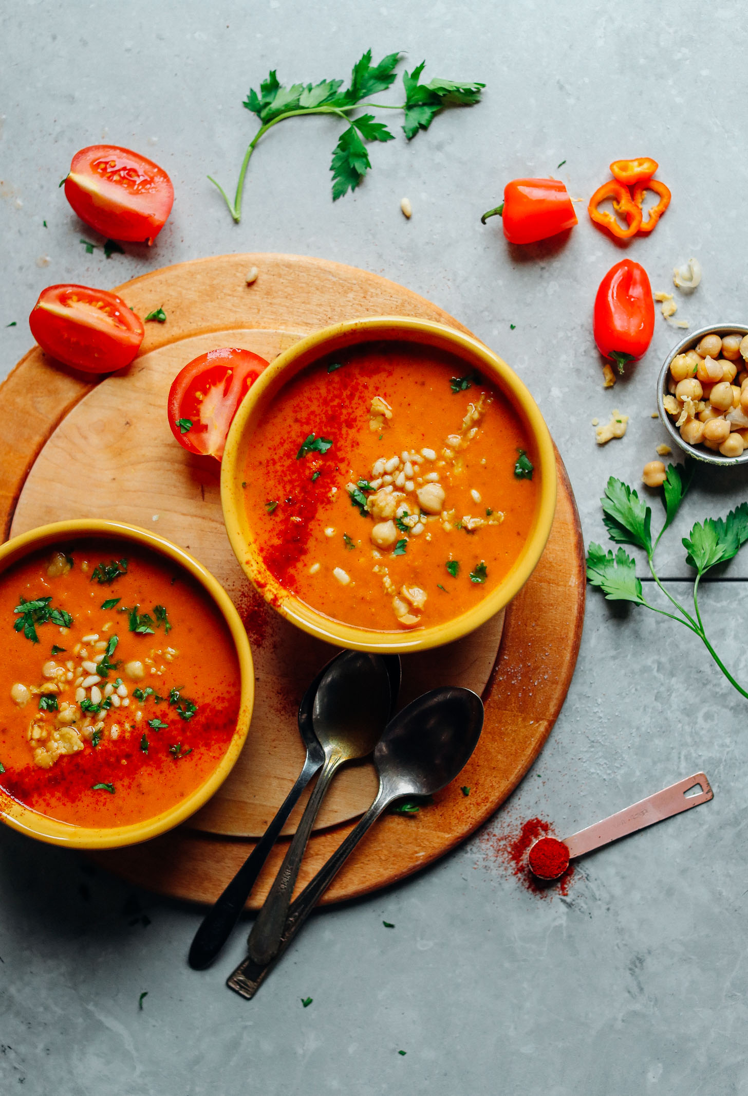 Two bowls of vegan Romesco Soup with Smashed Chickpeas surrounded by fresh tomatoes, red bell peppers, chickpeas, parsley, and smoked paprika