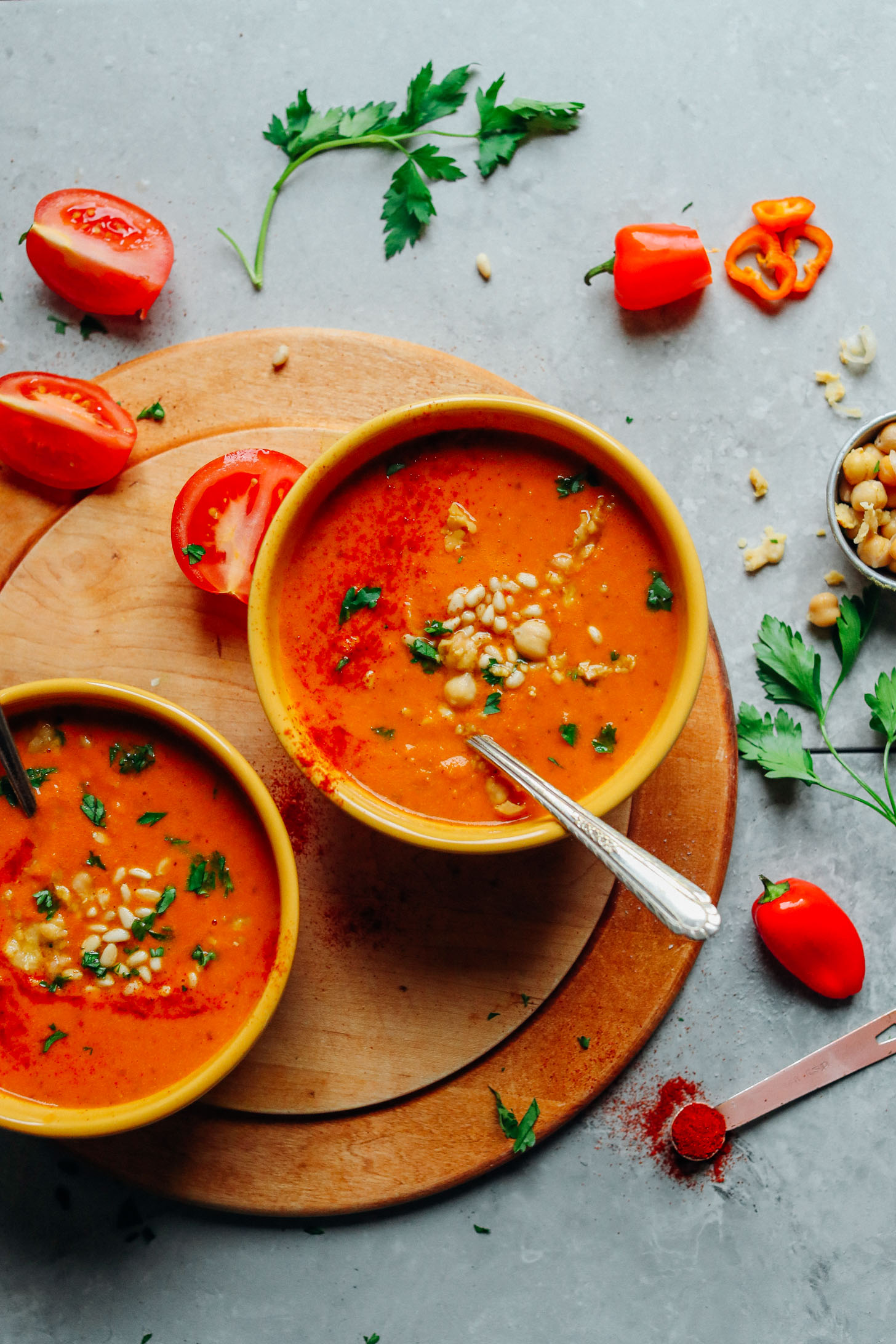 Two bowls of vegan Romesco Soup with Smashed Chickpeas surrounded by fresh tomatoes, peppers, parsley, smoked paprika, and chickpeas