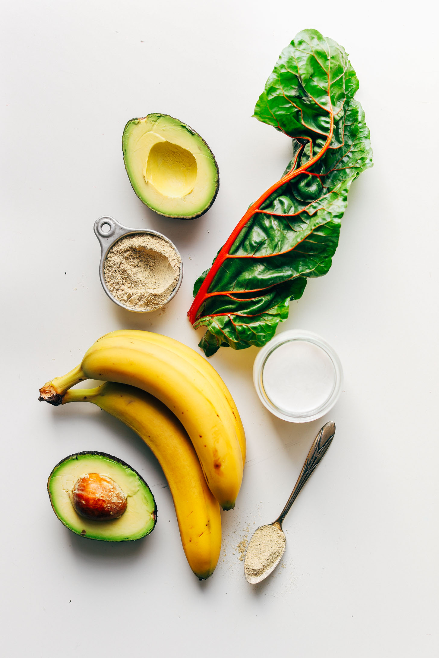 Avocado, bananas, chard, vanilla protein powder, almond milk, and maca laid out for making our Creamy Avocado Green Smoothie recipe