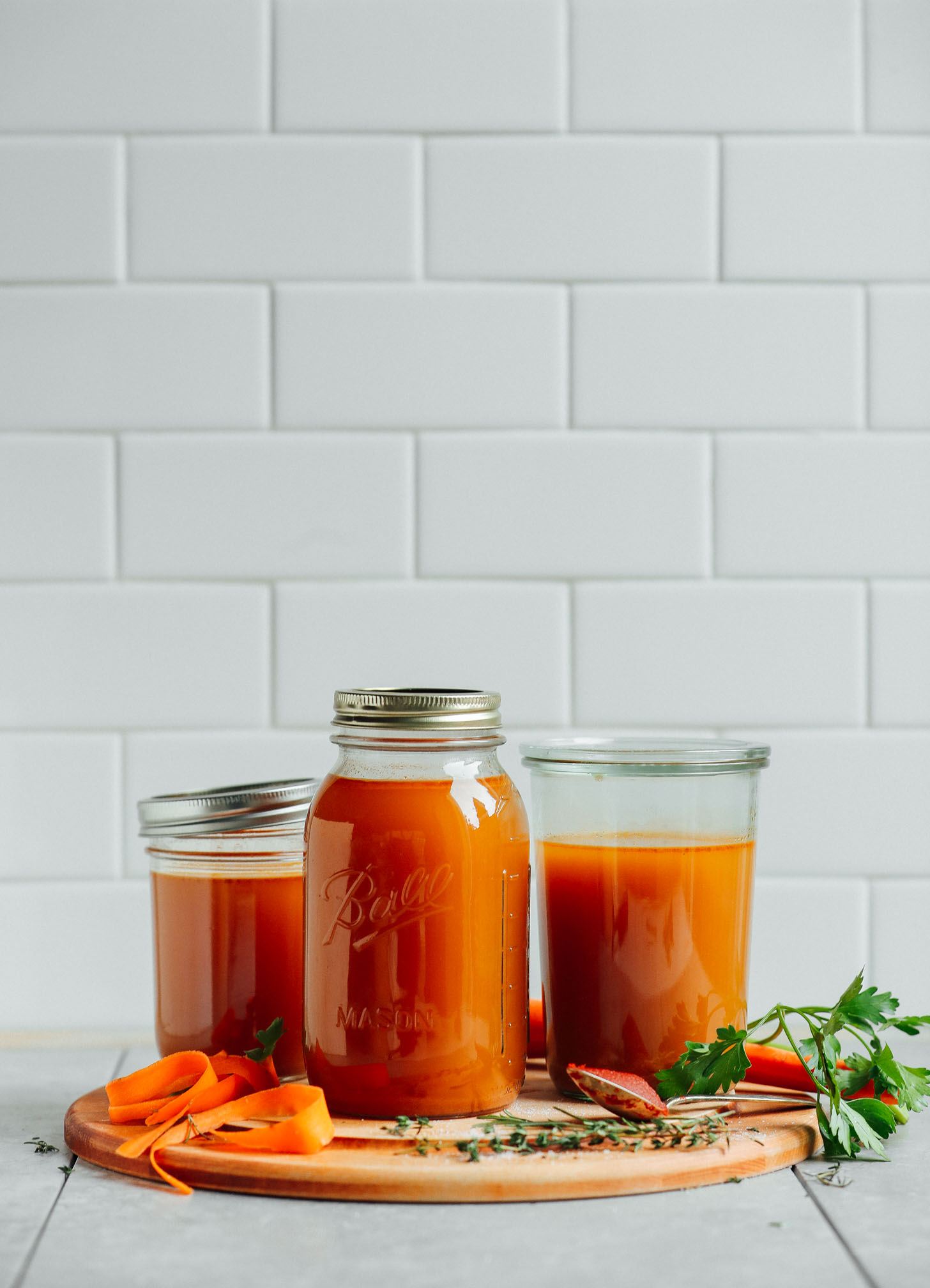 Three glass jars filed with the BEST homemade vegetable broth and surrounded by fresh carrots, thyme, parsley, and tomato paste