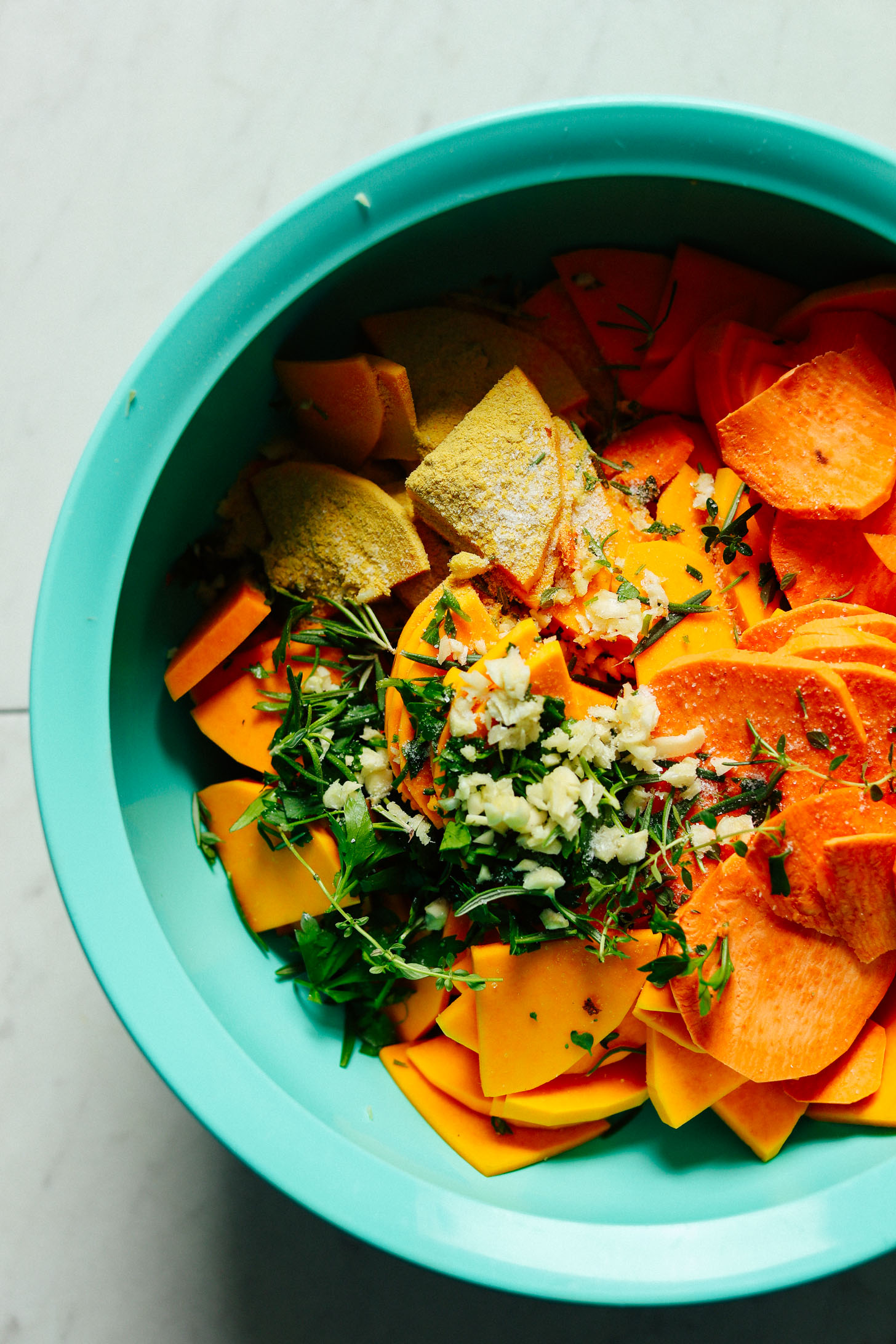 A big blue mixing bowl filled with thinly sliced squash and sweet potato, garlic, fresh herbs, and nutritional yeast