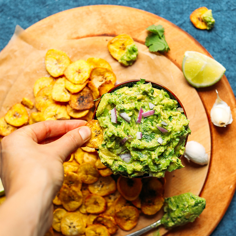 Using a homemade Baked Plantain Chip to scoop up Garlicky Guacamole from a bowl