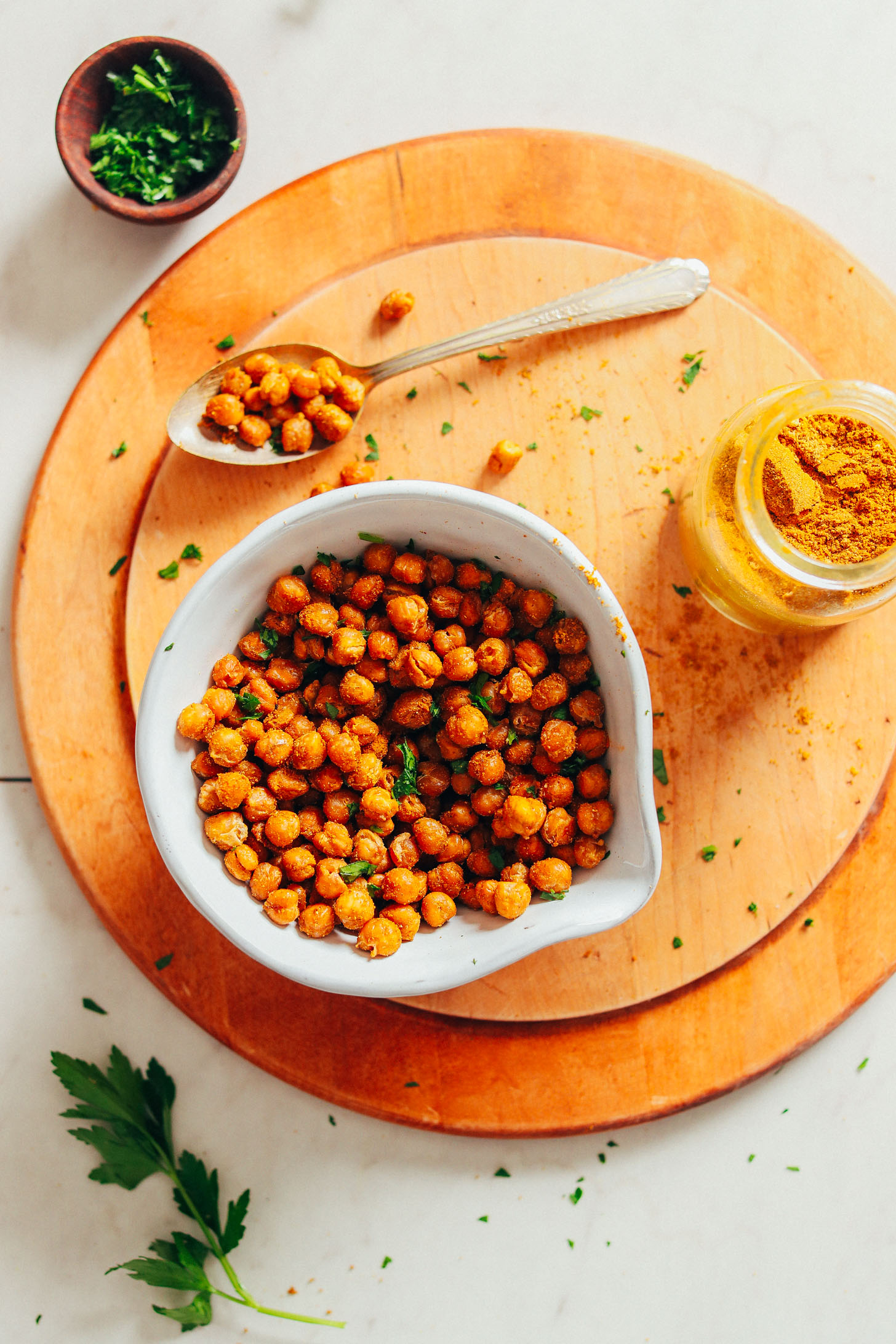 Wood cutting board with a bowl of Crispy Baked Chickpeas alongside fresh parsley, a spoonful of chickpeas, and curry powder
