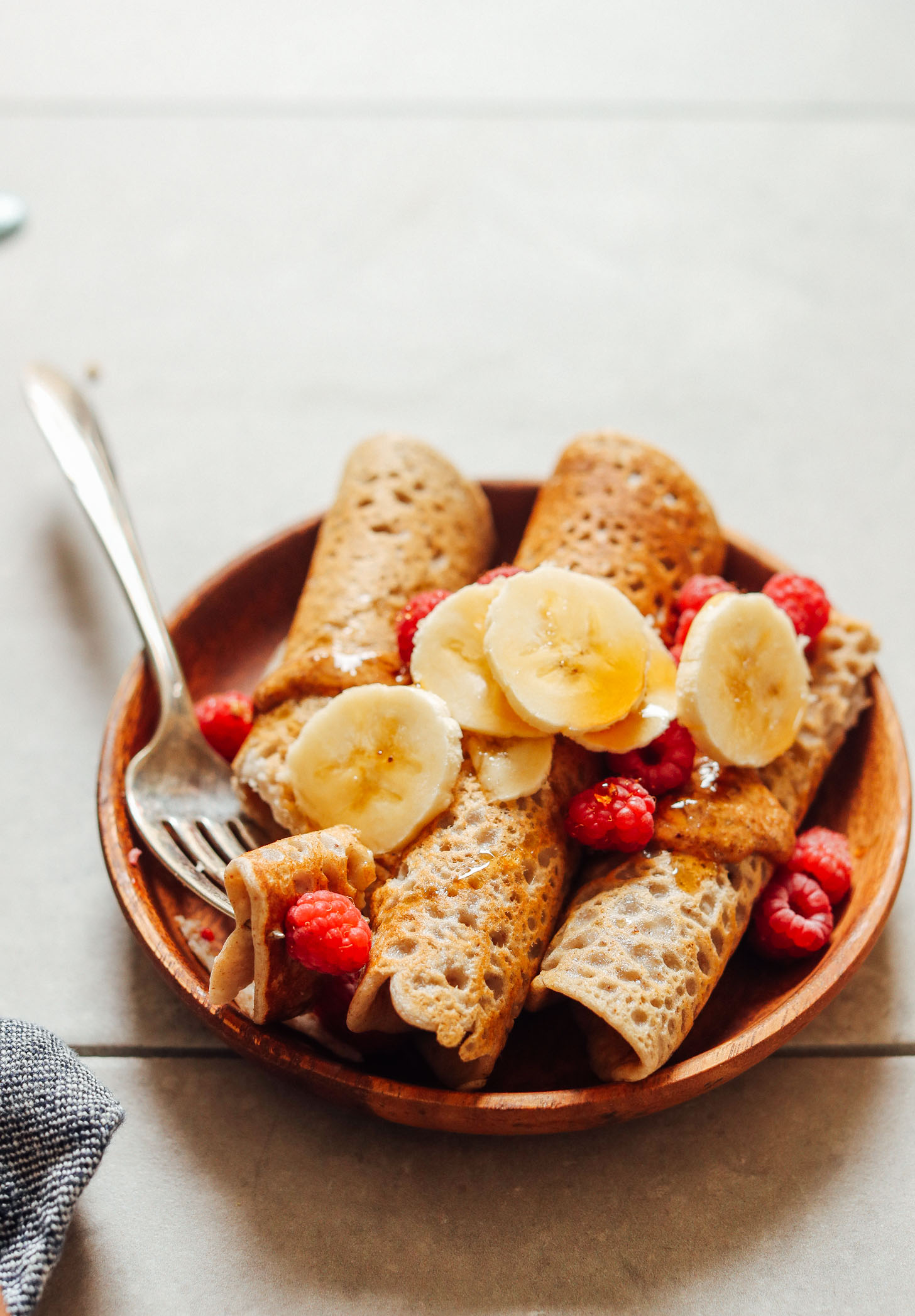 An eye level shot of buckwheat crepes on a wooden plate with bananas and raspberries