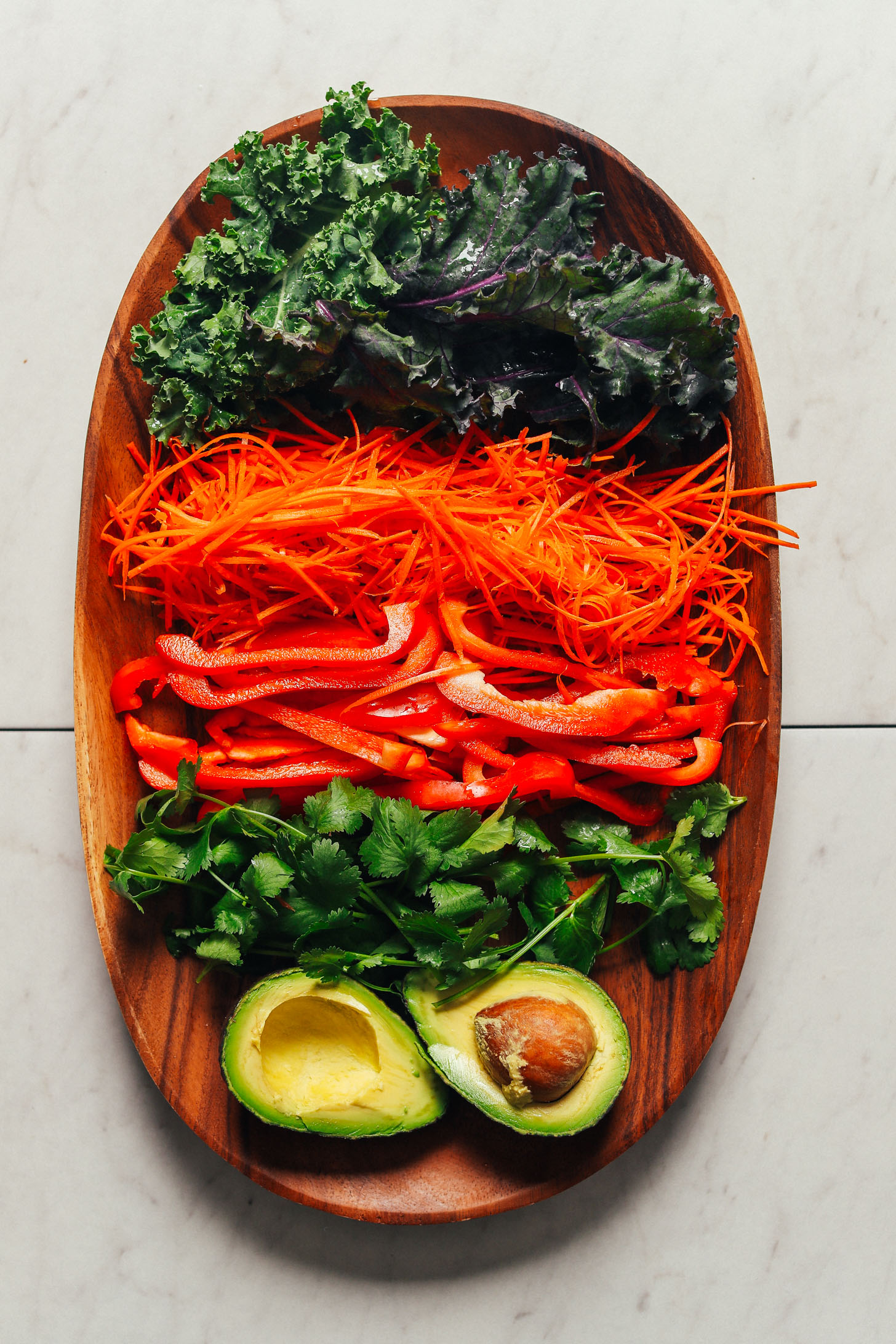 Platter of fresh vegetables including kale, shredded carrots, sliced red bell peppers, cilantro, and avocado